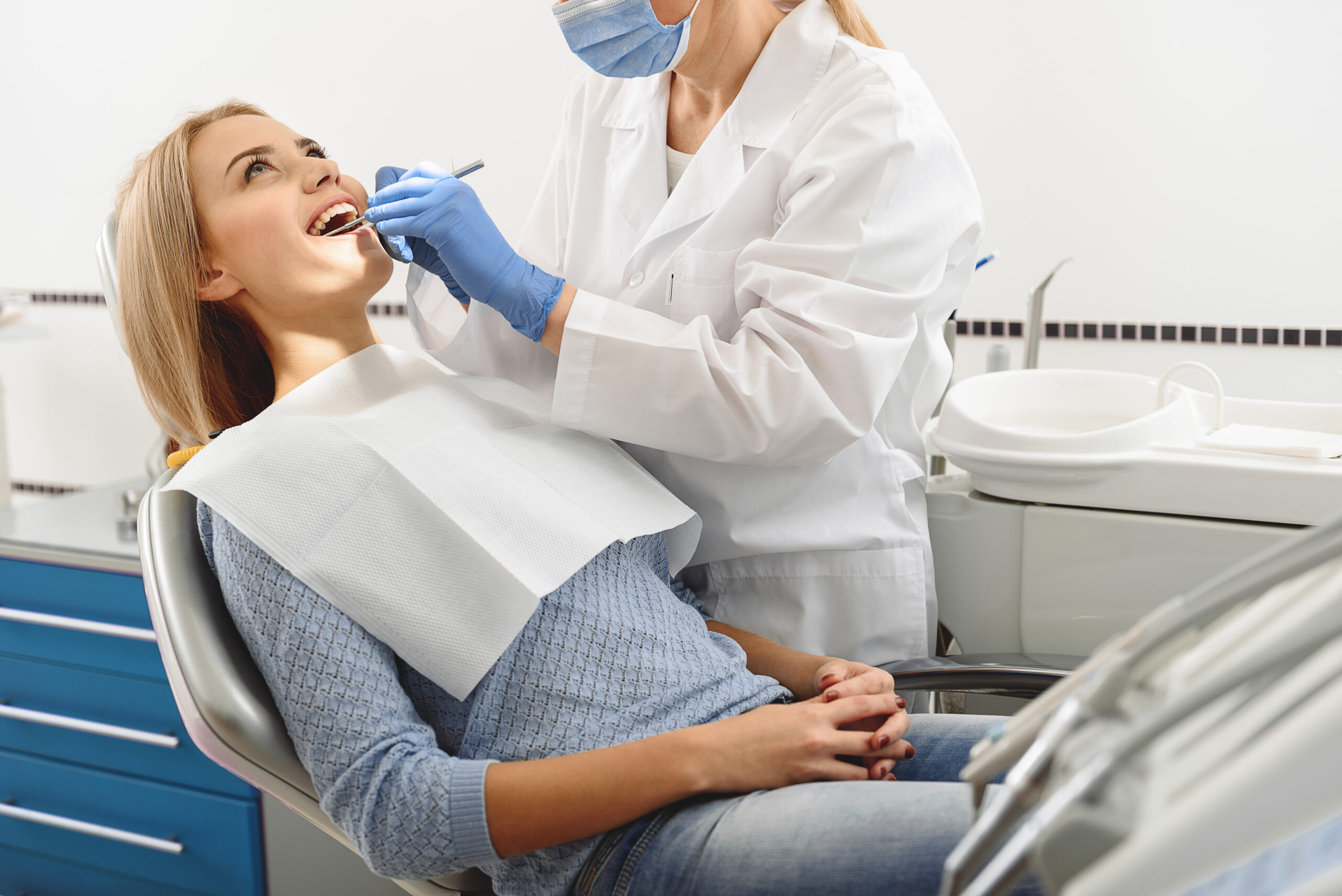 Dentist taking care of teeth of patient. Joyful women sitting with crossing arms on chair near standing doctor in bright dental room